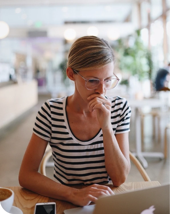 شخص جالس على طاولة مقهى، ينظر بتأمل إلى شاشة كمبيوتر محمول ويده قرب فمه، مما يوحي بالقلق أو اتخاذ القرار أثناء العمل عبر الإنترنت.